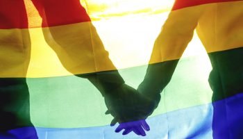 People, homosexuality, same-sex marriage, gay and love concept – close up of happy male gay couple hugging and holding rainbow flag