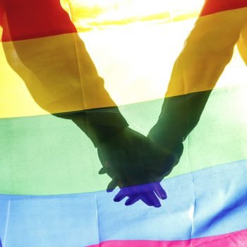 People, homosexuality, same-sex marriage, gay and love concept – close up of happy male gay couple hugging and holding rainbow flag